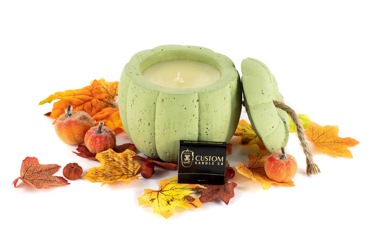 A Green Cement Candle with a lid, shaped like a pumpkin and surrounded by artificial autumn leaves and faux apples, sits next to a Custom Candle Co. label.