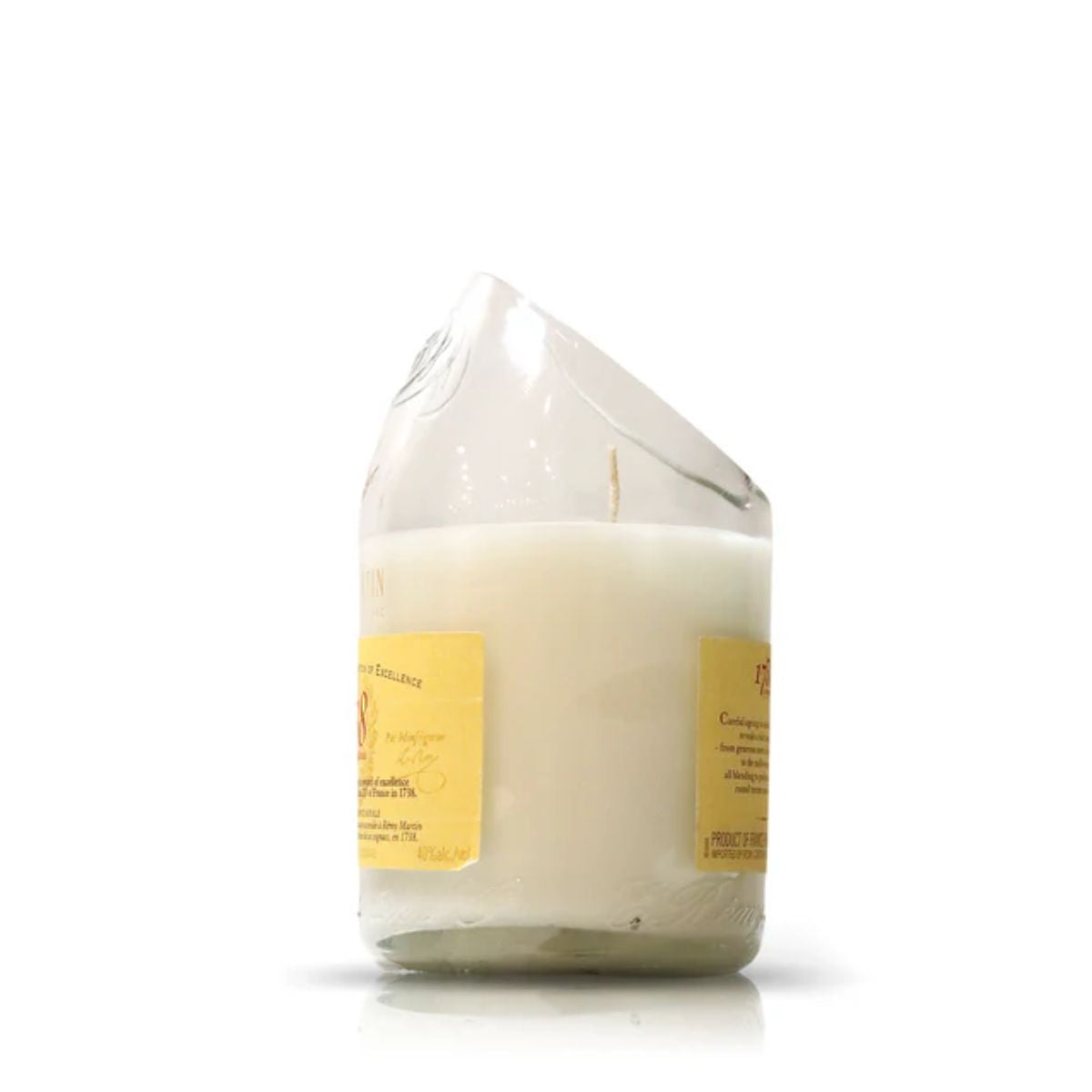 The Recycked 1738 Remy Martin Diagonal Cut Champagne Candle is a white artisanal candle in a clear, uniquely shaped glass with a yellow label, crafted from recycled bottles and shown against a plain white background.