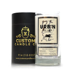 A Recycled Urbn De Luxe Cognac Candle made from clear glass sits next to a black and gold Custom Candle Co. cylindrical box.