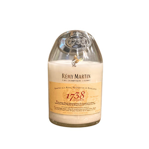 The Recycled Whiskey Candle, Remy 1738 features a clear cover, gold label, wax seal, and text honoring excellence. It's crafted from recycled materials with cruelty-free ingredients for an eco-friendly touch.