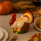 The Turkey Candle sits lit on a dining table surrounded by Thanksgiving décor, including a pumpkin and bread rolls in the background.