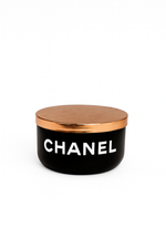 The Designer Noir Candle features a black round container with a metallic gold lid and "CHANEL" in white on the front, exuding elegance and sophistication.