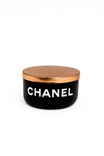 The Designer Noir Candle features a black round container with a metallic gold lid and "CHANEL" in white on the front, exuding elegance and sophistication.