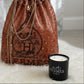 A brown studded Hermès drawstring bag with gold chains sits beside a black Designer Paris Candle on a white surface.