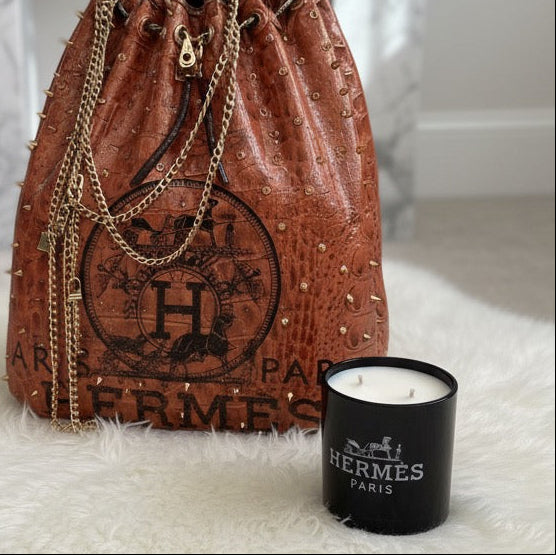 A brown studded Hermès drawstring bag with gold chains sits beside a black Designer Paris Candle on a white surface.