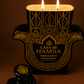 A black and gold Casa de Hamsa Añejo Tequila Candle, shaped like a Hamsa with two lit wicks and a matching stopper, rests on a brown background. Its label reads "Casa de Hamsa Tequila Añejo, Hecho en México.”.