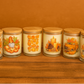 Six glass jars with wooden lids from the Fall Harvest Candle Collection feature autumn designs—sunflowers, pumpkins, candles, and a truck on a brown background—perfect for displaying your favorite hand-poured or Fall Harvest Candle.