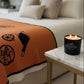 A Designer Paris Candle with coconut-apricot wax glows on a marble side table next to a neatly made bed, featuring an orange patterned blanket and beige pillows.