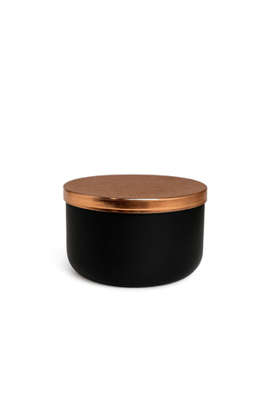 The Designer Noir Candle features a round black container with a metallic copper lid, adding sophistication to any space. Shown here against a plain white background.