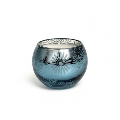 The Metallic Snowburst Candle features a blue glass holder with a floral metallic interior and round shape, making it perfect for holiday decor. Displayed on a white background for an elegant touch.