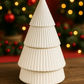 The Ceramic Tree Candle, a hand-poured white ceramic candle, rests on a wooden surface as both décor and keepsake, with blurred holiday lights twinkling in the background.