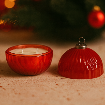 A Red Ornament Candle with a removable lid sits open on a surface, with a blurred Christmas tree in the background.
