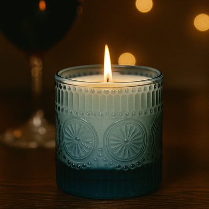 The Blue Embossed Glass Candle, made from natural wax and presented in a decorative blue glass holder, sits on a wooden surface with blurred warm lights and a wine glass in the background.