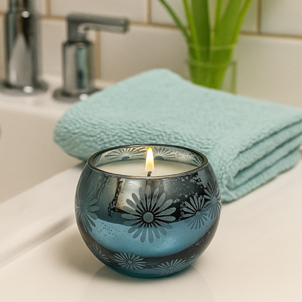 The Metallic Snowburst Candle, in a blue glass holder with floral designs, is displayed on a bathroom counter beside a turquoise towel and a green plant.