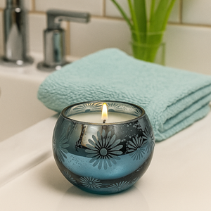 The Metallic Snowburst Candle, in a blue glass holder with floral designs, is displayed on a bathroom counter beside a turquoise towel and a green plant.