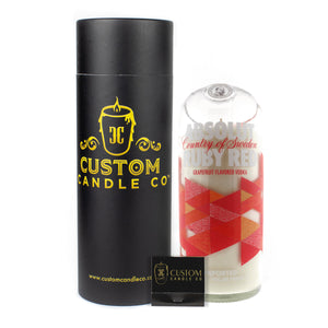 The Recycled Vodka Candle – Absolut Red Ruby, hand-poured in a glass shaped like an Absolut bottle, sits beside a black Custom Candle Co. tube and matchbox for a unique gift.
