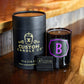 A black cruelty-free "Recycled Champagne Candle – Luc Belaire Rose Fantome" labeled with a purple “B” sits beside a black box, a matchbox, and a gold and black "Custom Candle Co." container on a wooden surface.