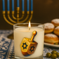 A Dreidel Candle, hand-poured in glass with a dreidel motif, sits on a patterned cloth; a menorah and donuts add festive Hanukkah ambiance in the background.