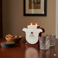 A Casa de Hamsa Reposado Tequila Candle, two empty crystal glasses, cinnamon sticks in a bowl, and coasters are arranged on a wooden table in a softly lit room.