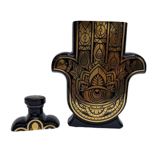 Casa de Hamsa Añejo is a black and gold perfume bottle shaped like a Hamsa hand, featuring intricate designs and an eye motif. Display it with its matching cap—ideal for pairing with a custom candle or all-natural soy decor.