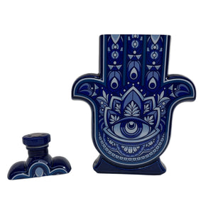 The Casa de Hamsa Blanco is a ceramic container shaped like a Hamsa hand, featuring a blue and white decorative pattern, shown with its matching lid beside it on a white surface.