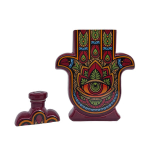 The Casa de Hamsa Reposado features a decorative, hamsa-shaped soy candle bottle with a vibrant eye and floral design, shown with its matching cap on a white background.