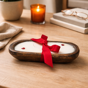The Red Ribbon - Dough Bowl Candle, featuring a rustic wooden design with red ribbon and heart accents, sits beside a lit candle, books, glasses, and a potted plant to bring charm to your cozy space.