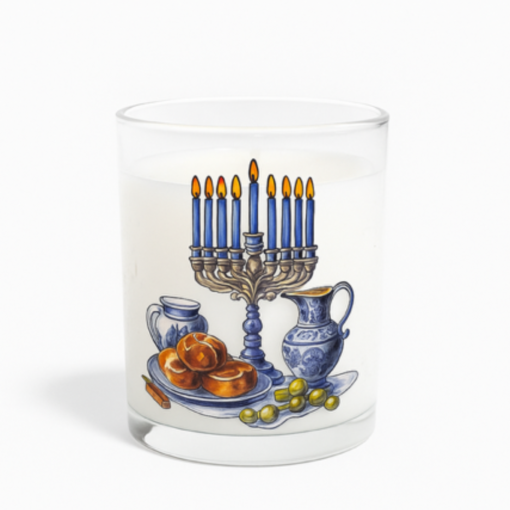 The Hanukkiah Candle is a white, hand-poured candle in a glass holder with an illustration of a lit Hanukkiah, challah bread, grapes, and blue ceramic pitchers—perfect festive décor for Hanukkah.