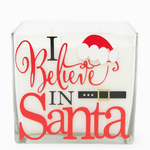 The 4-Wick “I Believe in Santa” Candle features white wax in a glass holder with festive red and black text, plus a Santa hat and belt graphic—perfect for adding Christmas cheer to your holiday décor.
