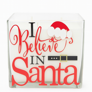 The 4-Wick “I Believe in Santa” Candle features white wax in a glass holder with festive red and black text, plus a Santa hat and belt graphic—perfect for adding Christmas cheer to your holiday décor.