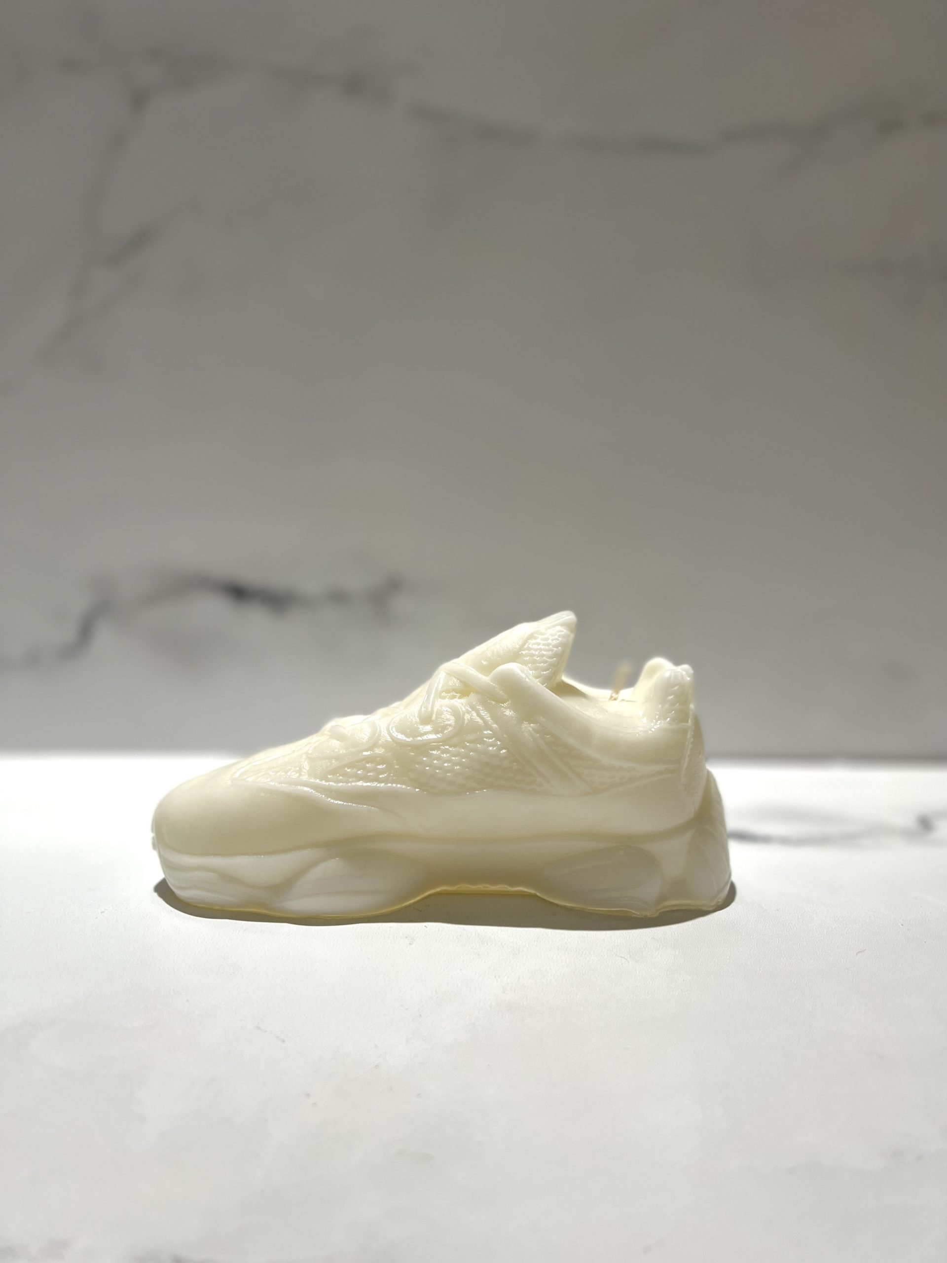 The Y500 – White Sneaker Candle – Small, shaped like a classic sneaker, sits on a white surface with a light gray marbled background. This hand-poured candle offers the refreshing black ice fragrance.