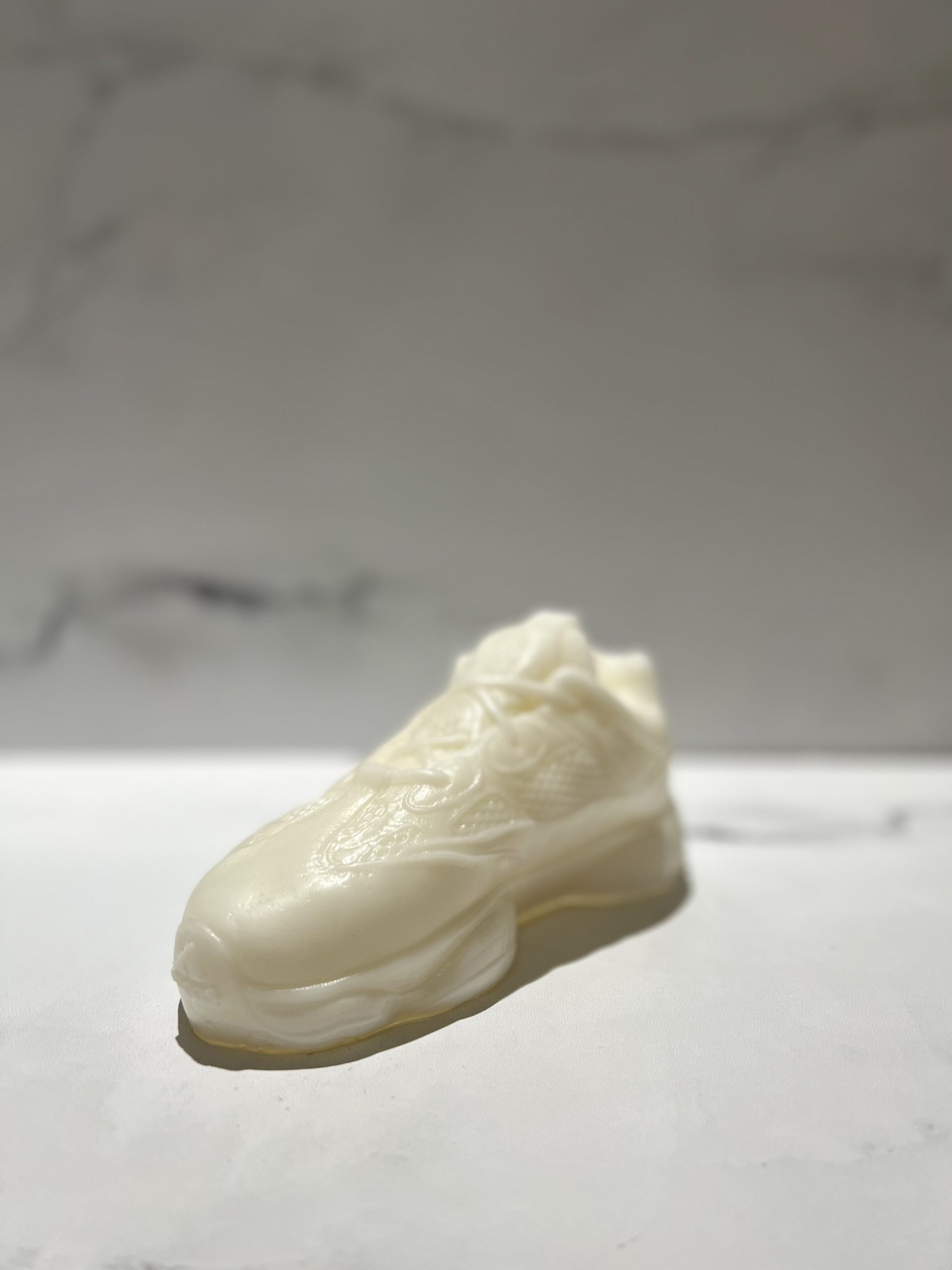 The Y500 – White Sneaker Candle – Small is a hand-poured, white sneaker-shaped candle, possibly black ice scented, displayed on a white surface with a marble-patterned background.