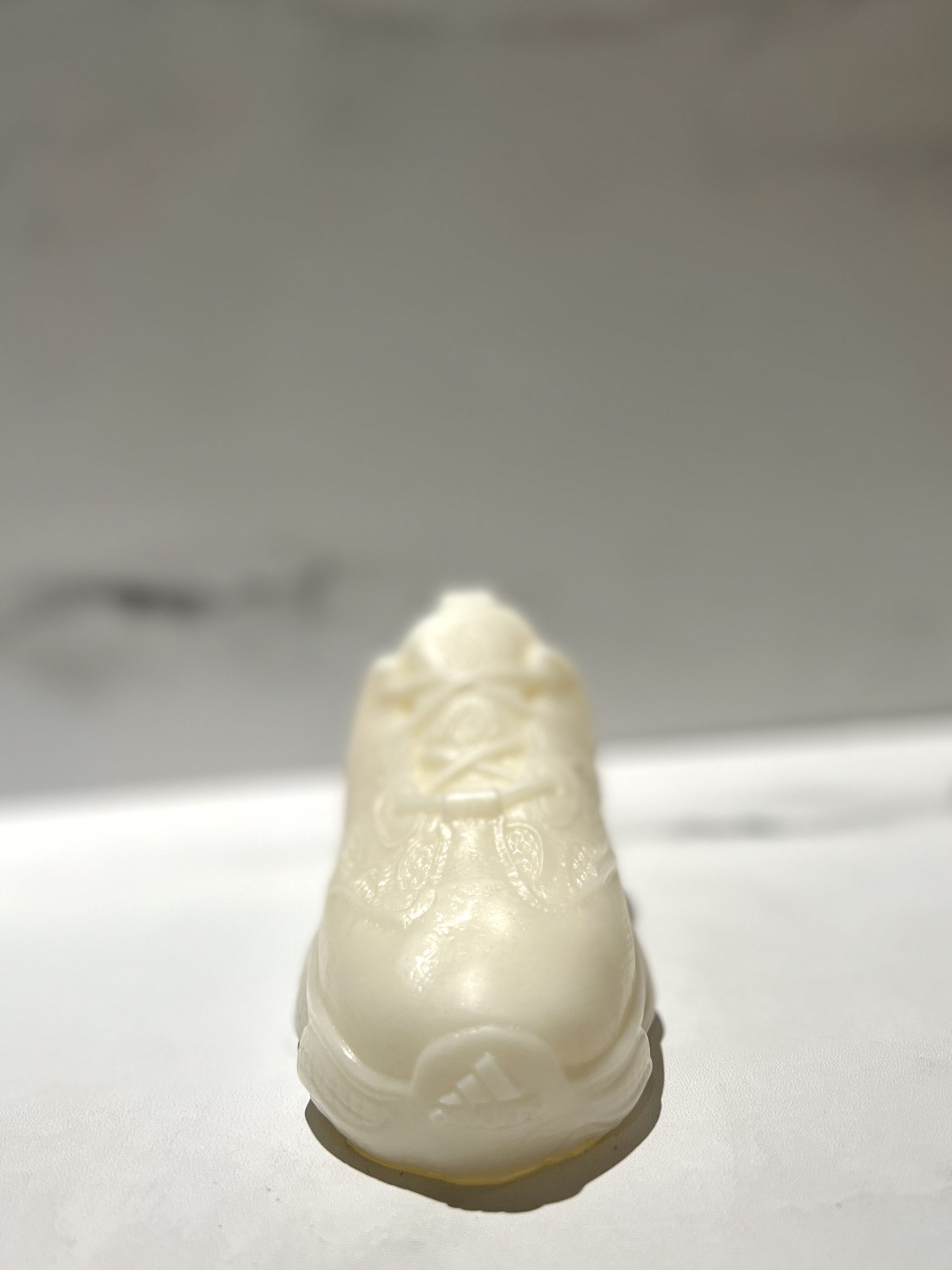 The Y500 – White Sneaker Candle – Small is a hand-poured white wax candle shaped like a sneaker, resting on a smooth white surface with a blurred light background and featuring a subtle black ice fragrance.