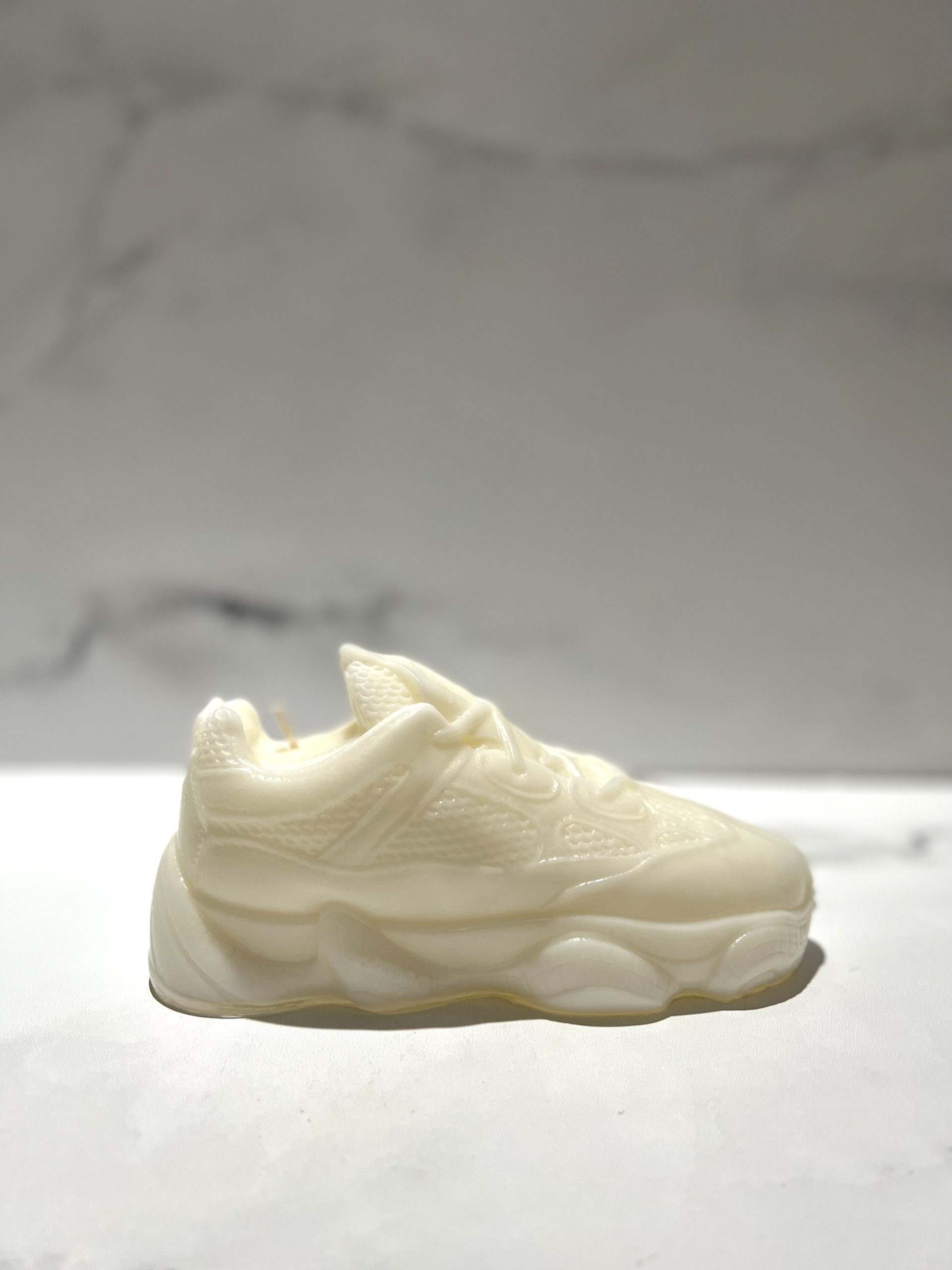 The Y500 – White Sneaker Candle – Small is a hand-poured candle shaped like a white sneaker, displayed on a light surface with a softly blurred background.