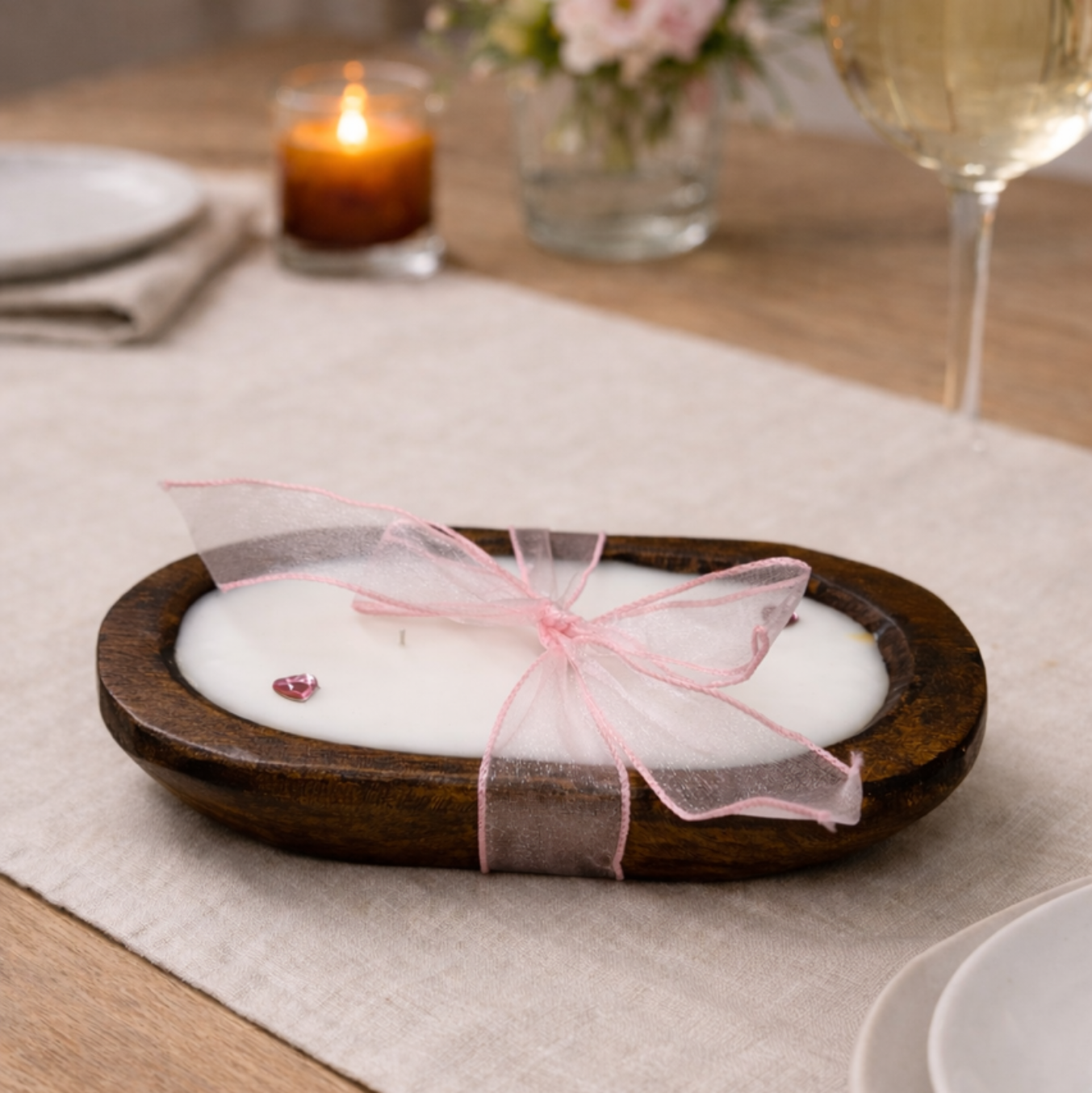 The Pink Ribbon - Dough Bowl Candle, a hand-poured soy candle in a brown oval wooden holder with a sheer pink ribbon, sits on a beige tablecloth beside plates, a wine glass, and another lit candle.