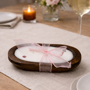 The Pink Ribbon - Dough Bowl Candle, a hand-poured soy candle in a brown oval wooden holder with a sheer pink ribbon, sits on a beige tablecloth beside plates, a wine glass, and another lit candle.