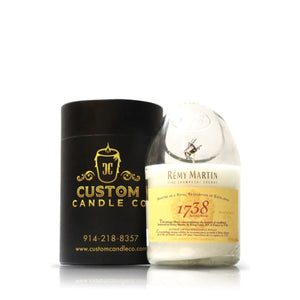The Recycked 1738 Remy Martin Diagonal Cut Champagne Candle sits beside a black cylindrical Custom Candle Co. container with gold lettering, highlighting the unique charm of recycled bottle candles.