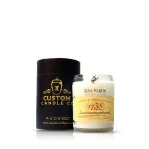 The Recycled Remy Martin Champagne Cognac Candle, in a glass with the "Rémy Martin 1738" label, sits beside a black Custom Candle Co. container, highlighting an eco-friendly, elegant use of recycled bottles.