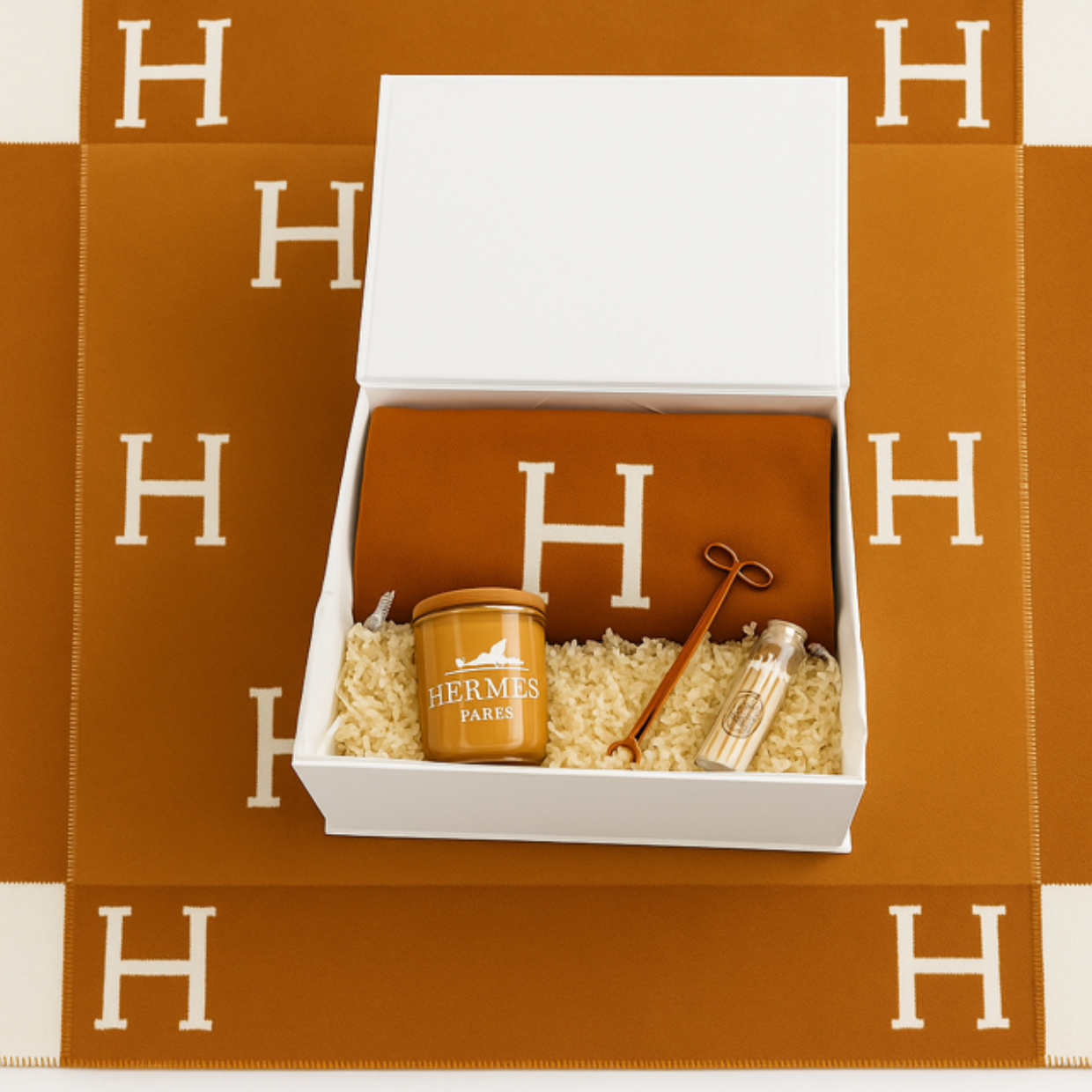 The Designer Orange Paris Gift Set includes a cotton throw with a white "H", candle, small bottle, and stirring stick, all elegantly arranged on an orange Hermes-patterned mat in an open white gift box.