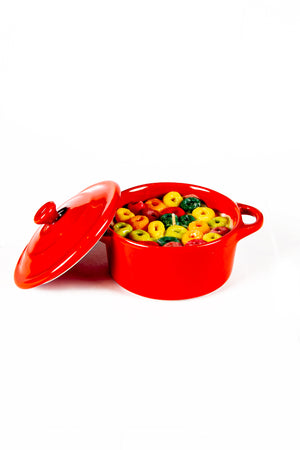 The Red Mini Cereal Crockpot Candle features a bright red casserole dish with a partially open lid, filled with colorful ring-shaped cereal pieces and milk, set against a white background.