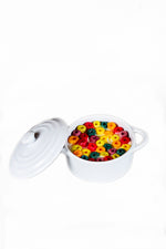 The White Mini Cereal Crockpot Candle sits with its lid off, filled with colorful ring-shaped cereal pieces, all set against a white background.