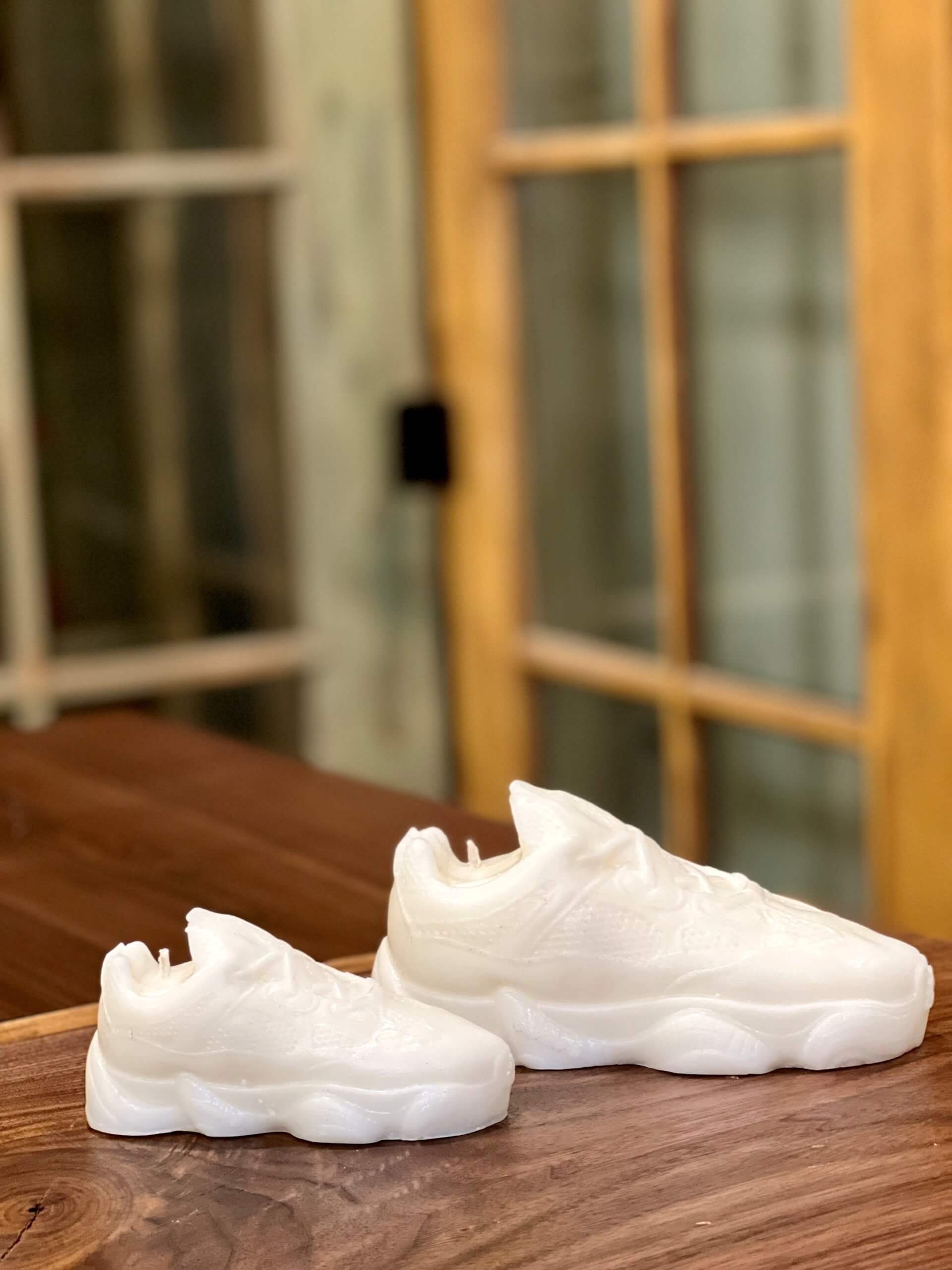 Two Y500 – White Sneaker Candle – Small, each meticulously hand poured, are displayed on a wooden surface before glass-paneled wooden doors.