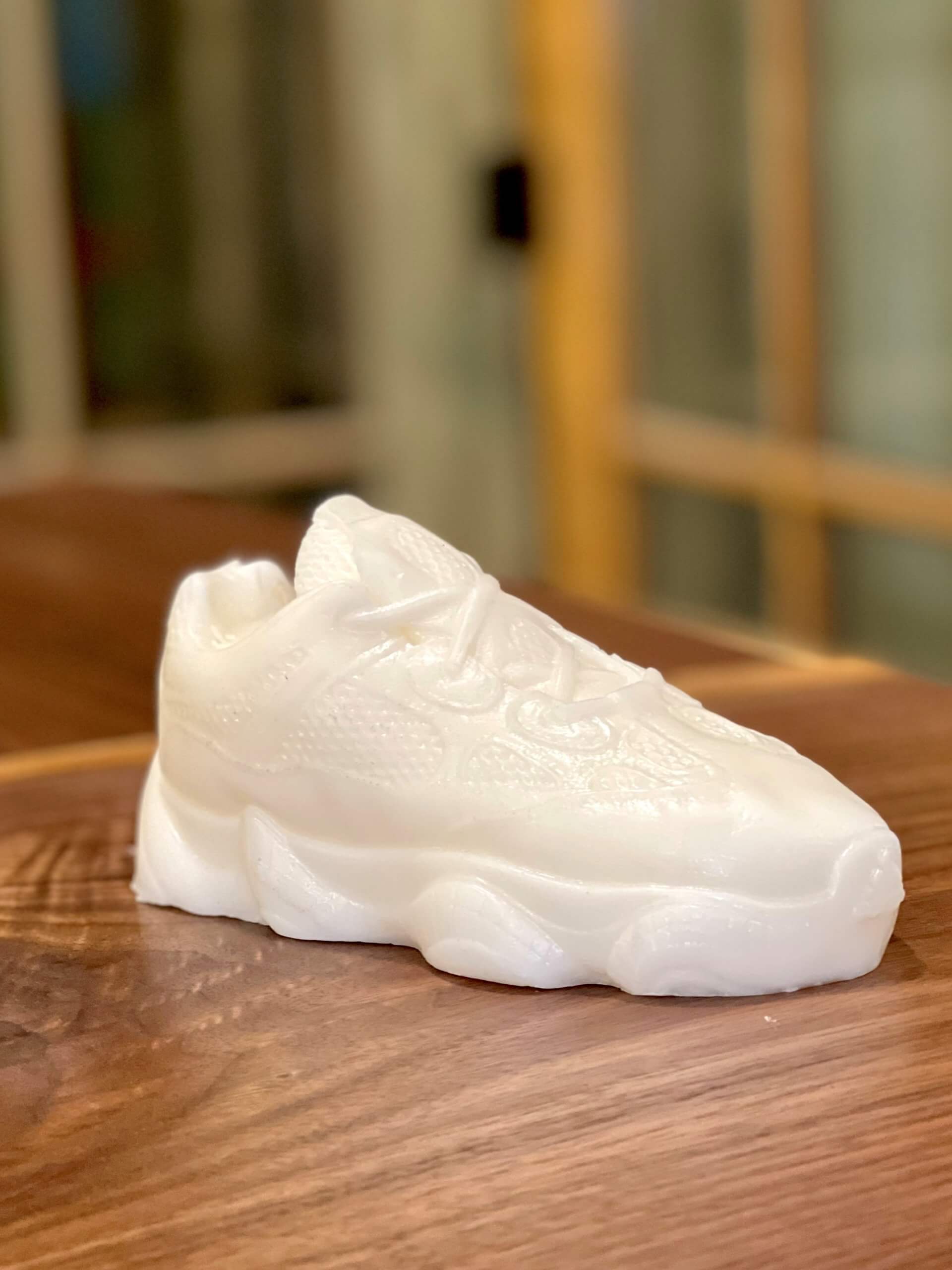 The Y500 – White Sneaker Candle – Small, hand-poured and wax-like, sits on a wooden surface—perfect for adding unique style and a subtle fragrance to any space.
