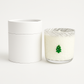 The Christmas Tree Glass Candle is a hand-poured candle in a glass holder with a small green Christmas tree decoration, displayed beside a round white container.