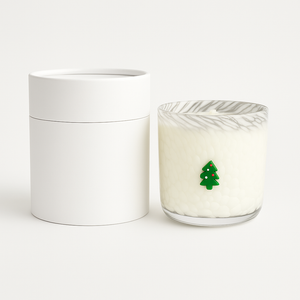 The Christmas Tree Glass Candle is a hand-poured candle in a glass holder with a small green Christmas tree decoration, displayed beside a round white container.
