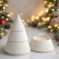 The Ceramic Tree Candle features white, ribbed ceramic containers stacked in a cone shape to mimic a Christmas tree, with one open to reveal a hand-poured candle—perfect for minimalist holiday decor amidst festive lights and greenery.