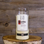 The Recycled Imported Vodka Candle, made from a repurposed vodka bottle and natural soy wax, rests on a wooden surface with a rustic wood backdrop.