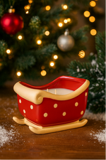 The Sleigh Candle, a red ceramic candle with gold polka dots, sits on a wooden surface. Festive decor and twinkling lights in the background enhance its warm holiday glow, making it a charming addition to your Christmas setting.
