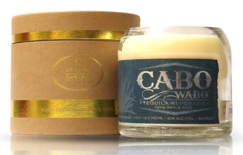A candle with a gold box next to it featuring Recycled Cabo Wabo Reposado Tequila.